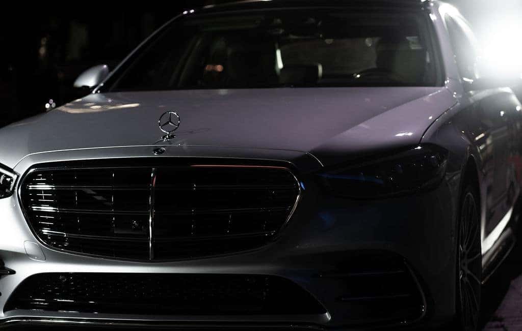 Front view of a shiny gray Mercedes-Benz with a prominent grille and hood ornament, illuminated by bright lights.