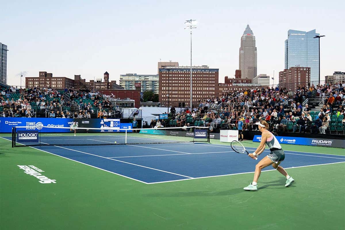 A female tennis player prepares to hit the ball on a blue and green court, with a city skyline and spectators in the background.