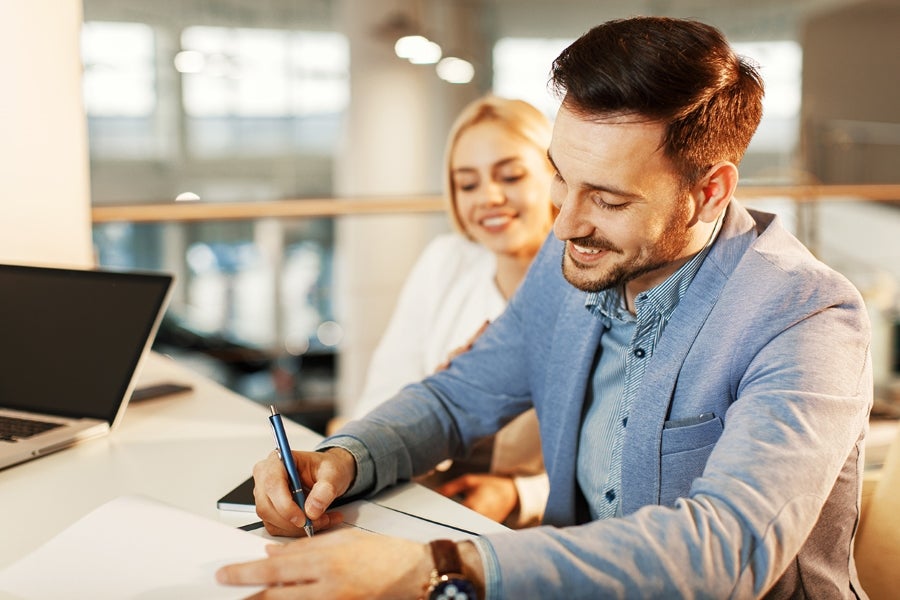 A smiling man signing paperwork with a woman looking on