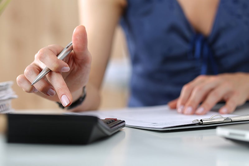 A person using a calculator while reviewing paperwork on a clipboard.