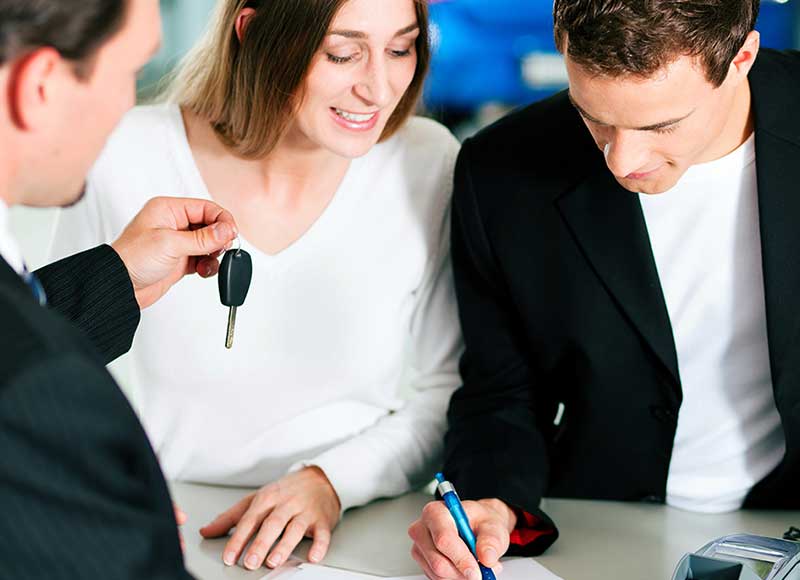 A couple signs paperwork while a car dealer holds out vehicle keys